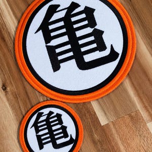 May include: Two circular embroidered patches featuring a white background with a black Japanese kanji character in the center. Each patch is bordered by a thick black ring and an outer orange stitched edge, creating a distinct contrast.
