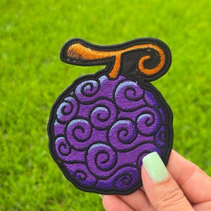 May include: An embroidered patch shaped like a round fruit with a dark purple surface covered in black swirling patterns and a thick black outline. The patch features a curved orange stem at the top, also outlined in black thread.