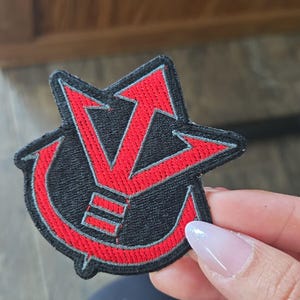 May include: Black and red embroidered patch with a stylized design. The patch features a black background with a red arrow-like symbol and a curved shape. The edges are outlined in red.