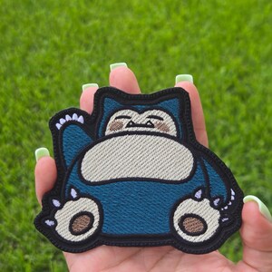 Peut inclure: Un &eacute;cusson brod&eacute; repr&eacute;sentant une cr&eacute;ature de dessin anim&eacute; avec une fourrure bleu fonc&eacute;, un ventre cr&egrave;me et des marques faciales beige. Le personnage a un corps arrondi, de petites oreilles et des griffes blanches, le tout bord&eacute; de fil noir.
