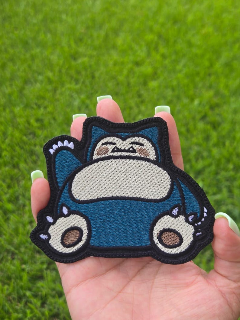 Peut inclure: Un &eacute;cusson brod&eacute; repr&eacute;sentant une cr&eacute;ature de dessin anim&eacute; avec une fourrure bleu fonc&eacute;, un ventre cr&egrave;me et des marques faciales beige. Le personnage a un corps arrondi, de petites oreilles et des griffes blanches, le tout bord&eacute; de fil noir.