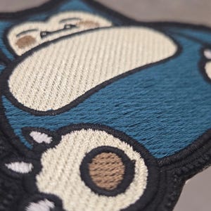 Peut inclure: Un &eacute;cusson brod&eacute; repr&eacute;sentant une cr&eacute;ature bleue stylis&eacute;e avec un ventre et un visage couleur cr&egrave;me. Le motif comprend une bordure noire, des griffes blanches et des accents circulaires marron sur les pieds, avec des d&eacute;tails de fils.