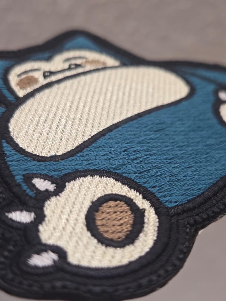 Peut inclure: Un &eacute;cusson brod&eacute; repr&eacute;sentant une cr&eacute;ature bleue stylis&eacute;e avec un ventre et un visage couleur cr&egrave;me. Le motif comprend une bordure noire, des griffes blanches et des accents circulaires marron sur les pieds, avec des d&eacute;tails de fils.