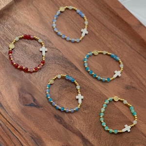 May include: Five beaded bracelets with gold accents and white cross charms are displayed on a wooden tray. The bracelets feature various colors of beads, including red, blue, and green. Each bracelet has a gold medallion.