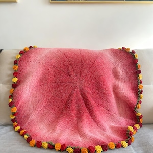 May include: A handmade, round blanket in shades of red and pink, with a floral border of yellow, orange, and red crocheted roses. The blanket is draped over a light-colored sofa, showcasing its soft texture and intricate design.