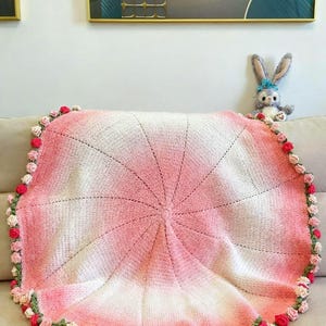 May include: A round, crocheted blanket with a gradient of white to pink hues. The blanket is edged with small, crocheted roses in shades of pink and red, with green leaves. A plush bunny toy with a blue bow sits nearby.