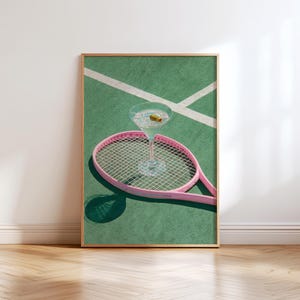 May include: A framed print featuring a martini glass with an olive resting on a pink tennis racket. The racket is positioned on a green tennis court with white lines. The image has a retro, sporty aesthetic.