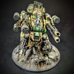 May include: A detailed miniature war machine, painted in olive green, tan, and metallic silver. The model features multiple weapon systems, including missile launchers and cannons, and is mounted on a textured base. The design incorporates intricate mechanical details and glowing green accents.
