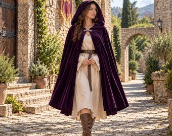 May include: A deep purple velvet hooded cloak featuring a silver-toned metal clasp at the neck, worn over a light beige long-sleeved dress with a brown leather belt and brown knee-high leather boots.
