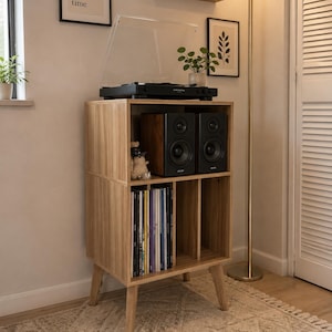 May include: A light wood record player stand with four tapered legs, featuring an open top shelf holding a black turntable and two black speakers, and a lower section with vertical dividers for vinyl record storage in a light oak finish.