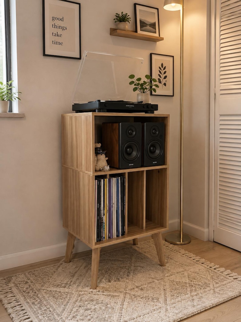May include: A light wood record player stand with four tapered legs, featuring an open top shelf holding a black turntable and two black speakers, and a lower section with vertical dividers for vinyl record storage in a light oak finish.