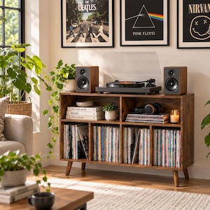 May include: A brown wooden media console with tapered legs featuring six storage compartments filled with vinyl records. The top surface holds a black turntable and two matching speakers. Three books titled The Vinyl Book, Music, and A Life in Sound sit on a shelf.