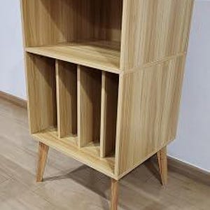 May include: A light wood grain storage unit featuring an open top shelf and a bottom section divided into four vertical compartments by three internal dividers. The piece is supported by four angled, tapered wooden legs in a matching light finish.