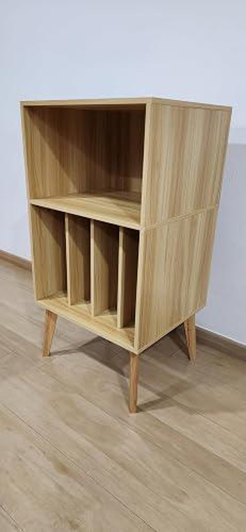 May include: A light wood grain storage unit featuring an open top shelf and a bottom section divided into four vertical compartments by three internal dividers. The piece is supported by four angled, tapered wooden legs in a matching light finish.