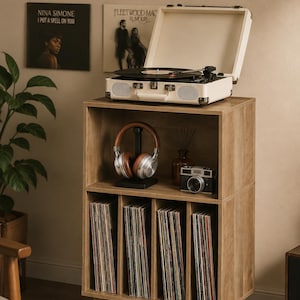 May include: A light wood record storage cabinet with four vertical dividers for vinyl records on the bottom shelf and an open top shelf holding a pair of brown and silver headphones and a vintage-style camera next to a cream-colored portable record player.
