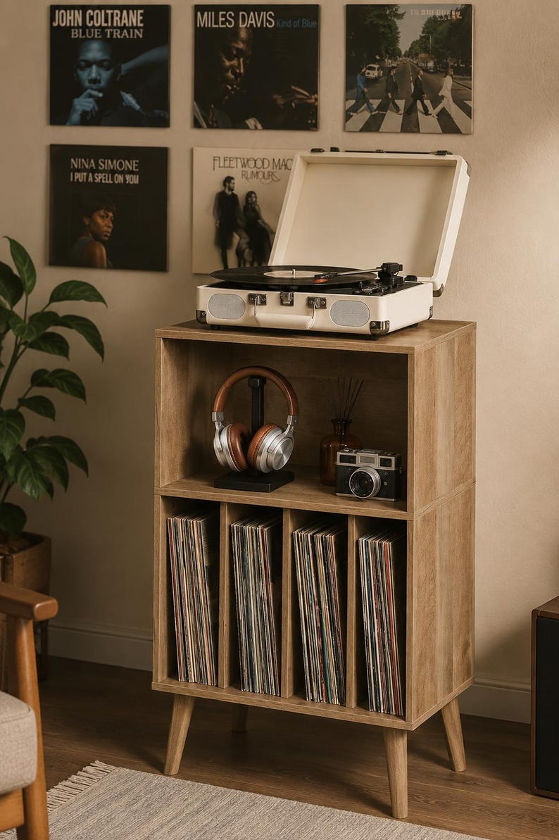 May include: A light wood record storage cabinet with four vertical dividers for vinyl records on the bottom shelf and an open top shelf holding a pair of brown and silver headphones and a vintage-style camera next to a cream-colored portable record player.