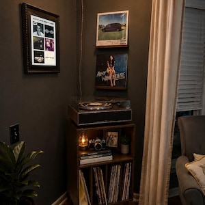 May include: A wooden record player console with a turntable on top and storage shelves below. The shelves hold a camera, books, and vinyl records. Two framed vinyl album covers, one by Amy Winehouse, are mounted on the dark wall above the console.