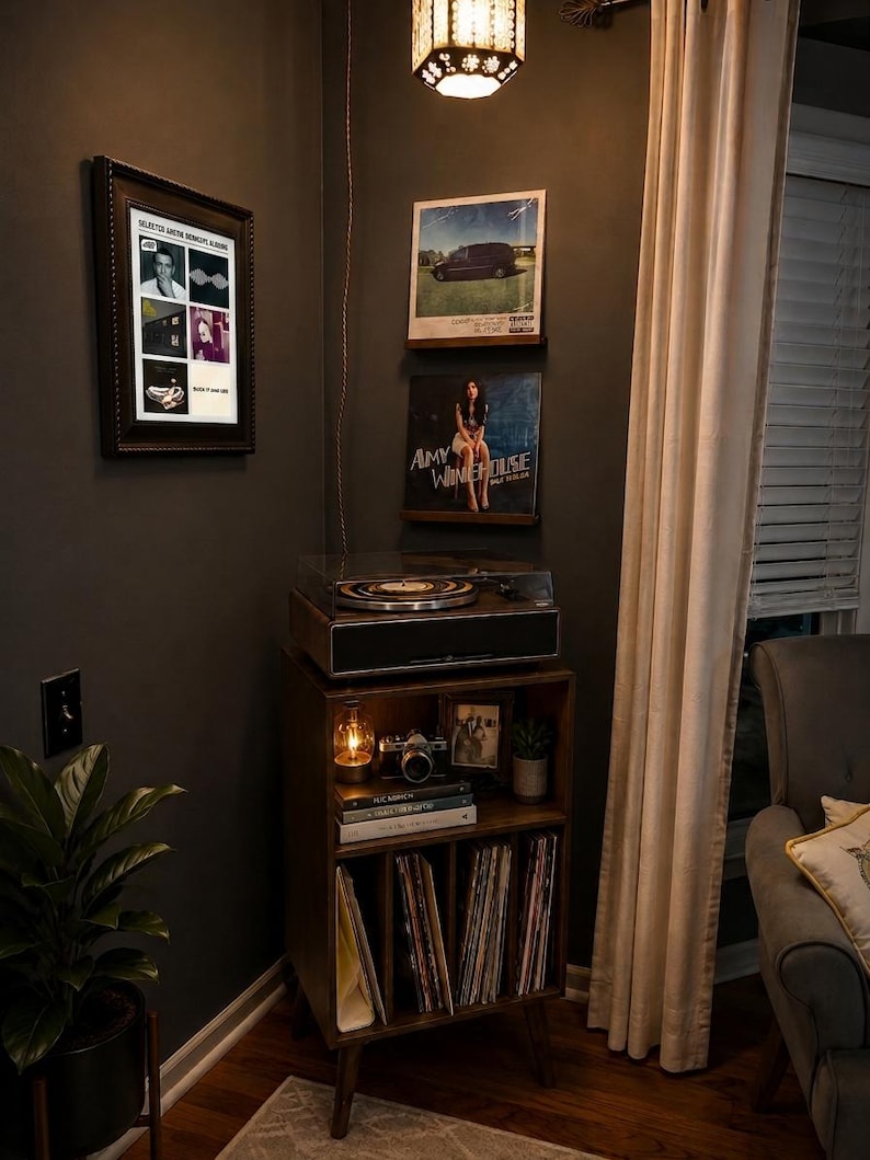 May include: A wooden record player console with a turntable on top and storage shelves below. The shelves hold a camera, books, and vinyl records. Two framed vinyl album covers, one by Amy Winehouse, are mounted on the dark wall above the console.
