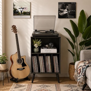 May include: A black wooden record player stand with a top surface holding a turntable, a middle shelf with books and headphones, and four lower vertical compartments filled with vinyl records, standing on four tapered legs in a room.