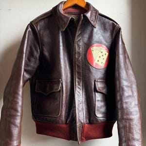 Op de afbeelding: Een bruine leren bomberjack met een rode gebreide kraag en manchetten. De jas heeft twee voorzakken en een ronde patch met speelkaarten op de borst. De jas hangt aan een houten hanger.