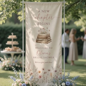 Puede incluir: Una pancarta vertical de tela color crema sobre un soporte de metal blanco con el texto A NEW CHAPTER BEGINS, WELCOME TO Olivia's BRIDAL SHOWER y We're so glad you're here, con una ilustración central de libros apilados y flores.