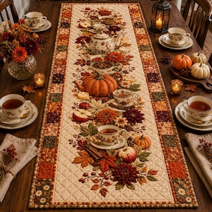 May include: A rectangular quilted table runner featuring a cream-coloured centre with an autumnal print of pumpkins, apples, cinnamon sticks, and flowers, bordered by a patchwork pattern of orange, brown, and green squares on a dark wooden table.
