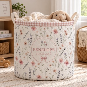 May include: A cylindrical quilted fabric storage basket featuring a floral pattern with pink and purple wildflowers, the name Penelope, and the text Penelope sweet girl inside a circular frame. It has two side handles and a pink gingham trim.