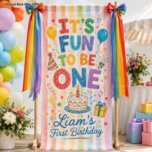 Op de afbeelding: Een verticale banner met pastelkleurige strepen met de tekst IT'S FUN TO BE ONE en Liam's First Birthday. Het ontwerp bevat illustraties van een verjaardagstaart, ballonnen, een feesthoedje en een cadeau, geaccentueerd door regenbooglinten.