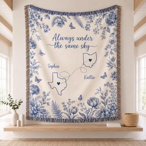 Op de afbeelding: Een crèmekleurige geweven deken met een blauwe bloemenrand, vlinders en de tekst Always under the same sky. Het toont de contouren van Texas en Ohio verbonden door een stippellijn, met de namen Sophia en Kallie erop gedrukt.
