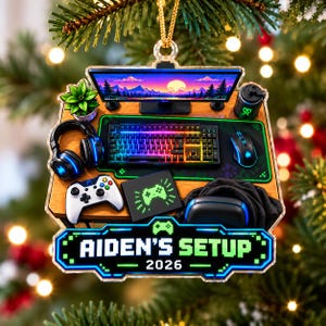 May include: A flat acrylic ornament featuring a gaming desk setup with a monitor, keyboard, mouse, controller, headphones, and a potted plant. A black sign at the bottom displays the text AIDEN'S SETUP 2026 in a green pixelated font.