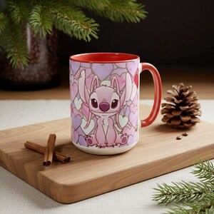 May include: A ceramic mug with a pink cartoon character and pink and purple hearts. The mug has a red interior and handle. The mug is on a wooden board with cinnamon sticks and a pine cone.