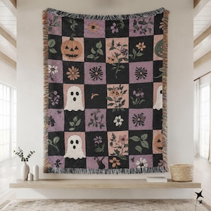 May include: A woven throw blanket featuring a checkered pattern of black and muted purple squares. The design includes illustrations of white ghosts, orange jack-o'-lanterns, and various floral and botanical motifs with fringed edges.