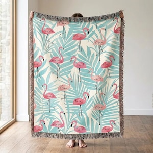 Op de afbeelding: Een geweven plaid met een herhalend patroon van roze flamingo's en lichtblauwe tropische palmbladeren op een crèmekleurige achtergrond. De deken heeft een textuurrijke franjerand aan de boven- en onderkant voor een decoratieve afwerking.