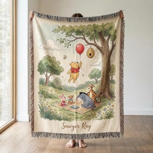 May include: A woven throw blanket featuring a colorful illustration of Winnie the Pooh characters in a forest setting. The design includes Pooh holding a red balloon, Piglet, Tigger, and Eeyore on a picnic blanket. The name Sawyer Ray is printed below.