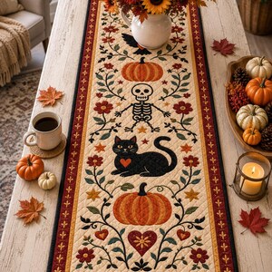 May include: A rectangular quilted table runner featuring a cream-coloured centre with a repeating pattern of orange pumpkins, a black cat with a red heart, a skeleton, and floral vines, bordered by a dark red and black frame with gold accents.