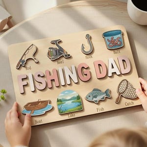 May include: Wooden fishing-themed puzzle with the words "FISHING DAD" in pink and white letters. The puzzle includes images of fishing gear, a boat, a lake scene, and a fish. The puzzle is held by a child.