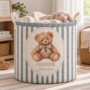 May include: A quilted fabric storage basket featuring blue and white vertical stripes and a central oval graphic of a brown teddy bear wearing a bow. The text OLIVER James is printed below the bear. The basket has two side handles and contains a plush toy.