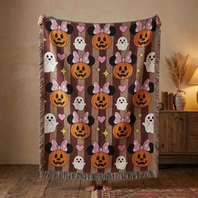 K&ouml;nnte beinhalten: Eine gewebte Wohndecke mit einem sich wiederholenden Muster aus orangefarbenen K&uuml;rbissen mit rosa Schleifen und schwarzen Mausohren, durchsetzt mit wei&szlig;en Geistern, kleinen rosa Herzen und gelben vierzackigen Sternen auf braun-beigem Streifengrund.