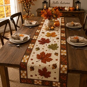 May include: A quilted table runner featuring a cream-coloured centre panel decorated with embroidered red, orange, and green maple leaves and brown acorns. The runner has a dark brown border with a patchwork pattern of small squares and leaf motifs.