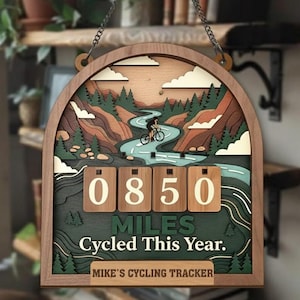May include: Wooden cycling tracker with a layered design. The arched frame surrounds a scenic illustration of a cyclist on a winding road. The tracker displays "0850 MILES CYCLED THIS YEAR" and "MIKE'S CYCLING TRACKER".