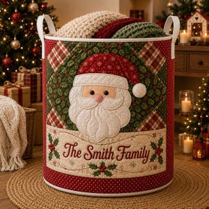 May include: A cylindrical fabric storage basket featuring a quilted Santa Claus face, a red and green holly pattern, and the text The Smith Family. The basket has two white handles and is filled with folded cream, red, and green knit blankets.