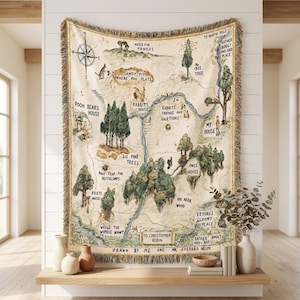 Puede incluir: Una manta tejida que muestra un mapa detallado del Bosque de los Cien Acres con ilustraciones de árboles, un río y una rosa de los vientos. El texto incluye etiquetas como Pooh Bear's House, Six Pine Trees y To Christopher Robin.