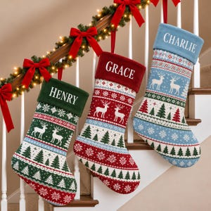 Peut inclure: Trois chaussettes de Noël personnalisées avec un motif tricoté de rennes, de sapins et de flocons de neige. La chaussette de gauche a un revers vert avec le nom HENRY, celle du centre un revers rouge avec GRACE, et celle de droite un revers bleu avec CHARLIE.