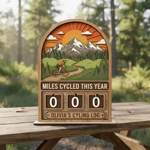 May include: Wooden cycling log with a mountain and sun design. The sign reads "MILES CYCLED THIS YEAR" and "OLIVIA'S CYCLING LOG". The counter displays "000". The background shows a cyclist on a path, trees, and mountains.