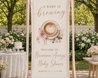 May include: A vertical cream-colored banner on a wooden stand featuring the text A BABY IS brewing, an illustration of a coffee cup with latte art, a pink ribbon, and the text WELCOME TO Brianna Avery's Baby Shower MARCH 15 2-5 PM.