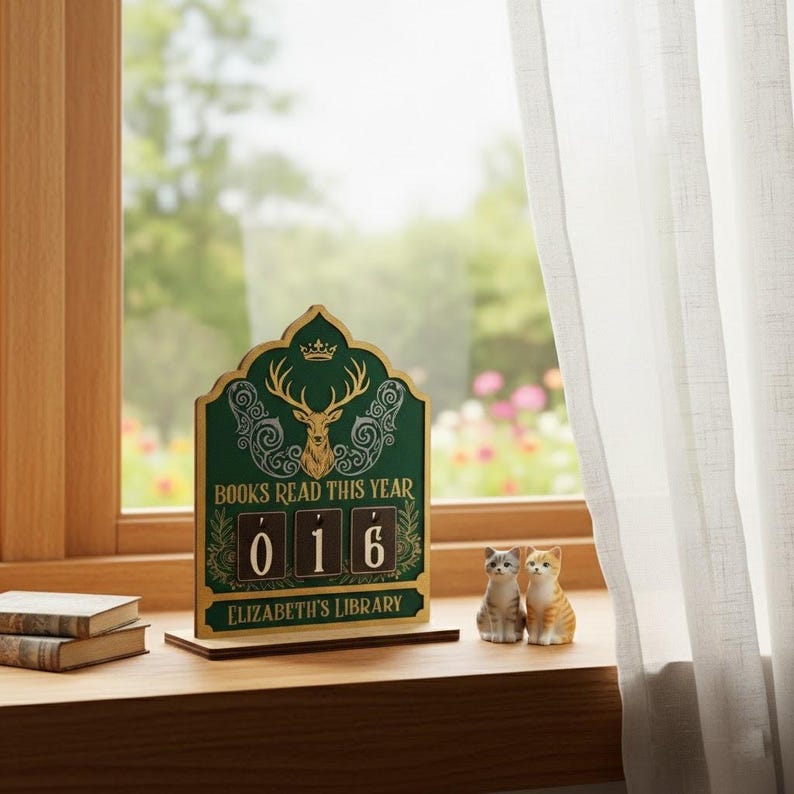 May include: A wooden book counter with a green background and gold detailing, showing "BOOKS READ THIS YEAR" and the number "016." The counter features a deer illustration and the text "ELIZABETH'S LIBRARY." Two small cat figurines are also present.