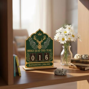 May include: A wooden desk calendar with a green and gold design. The calendar reads "BOOKS READ THIS YEAR" and displays "016". The design includes a deer head with antlers and the text "ELIZABETH'S LIBRARY". A vase of daisies and cat figurines are also visible.
