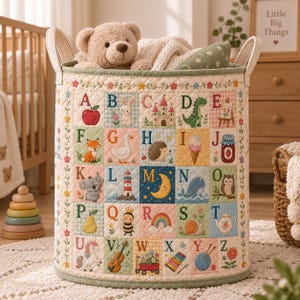 May include: A cylindrical quilted fabric storage basket featuring a colorful alphabet grid with embroidered illustrations for each letter from A to Z. A plush teddy bear sits inside the basket, which has two sturdy fabric handles on the sides.