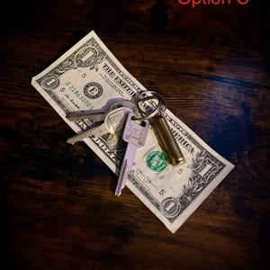 Peut inclure: Un jeu de clés est posé sur un billet d'un dollar, avec le texte "THE UNITED STATES OF AMERICA" visible. Les clés sont argentées et dorées, et le billet est incliné sur une surface en bois sombre. Le texte "Option C" est dans le coin supérieur droit.