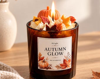 Könnte beinhalten: Eine brennende Kerze in einem braunen Glasgefäß mit einem Etikett, auf dem D.Light AUTUMN GLOW BIO-SOJAWACHS steht. Die Wachsoberfläche ist mit einem kleinen orangefarbenen Kürbis und verschiedenen herbstlichen Blattformen in Rot, Gelb und Orange verziert.